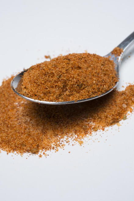 Fire & Smoke Society F&S | Pork Perfect Spice Rub 10.7 oz, close-up of a metal spoon filled with a reddish-brown spice rub, with some spilled on a white surface.