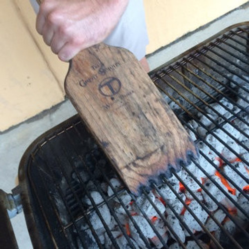 Great Scrape The Great Scrape Woody Paddle Grill Scraper, a wooden paddle grill scraper used for cleaning a barbecue grill, held above hot charcoal on the grill.