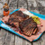 PK Grills Franklin BBQ Tray, a blue disposable food tray filled with sliced smoked brisket, pickles, onions, and BBQ sauce, placed on a rustic wooden surface.