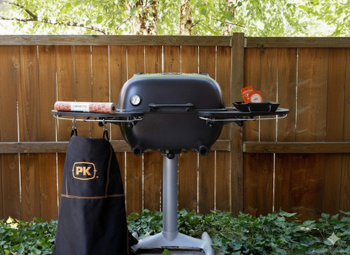 PK Grills PK360 Start Cookin' Bundle - Graphite, outdoor charcoal grill with side tray and cover, set up in a backyard with green trees and wooden fence.
