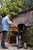 PK Grills The PK360 Grill & Smoker - Classic Silver, outdoor grill and smoker being used with flames visible, beside a wooden fence and greenery.