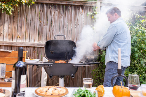 PK Grills The PK360 Grill & Smoker - Classic Silver, a black round grill and smoker with open lid releasing smoke, set outdoors with a man cooking, beside a wooden fence and a table with food and drinks.