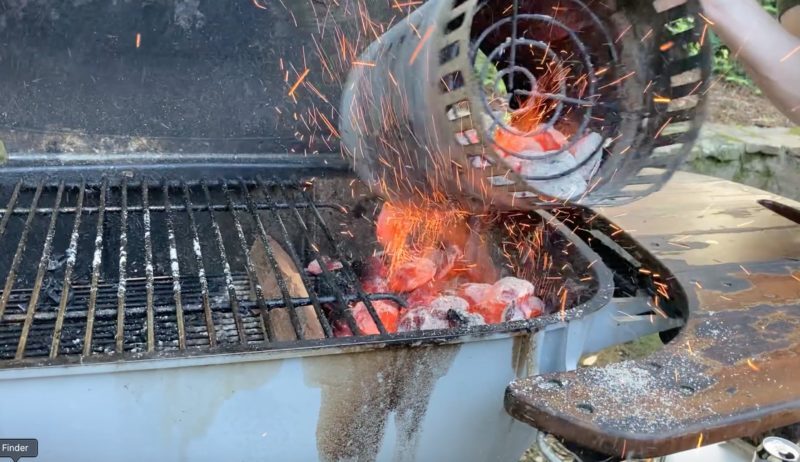 Glowing coals pouring from a metal chimney starter into a PK charcoal grill, sparks flying as hot briquettes fall into the cooking chamber.