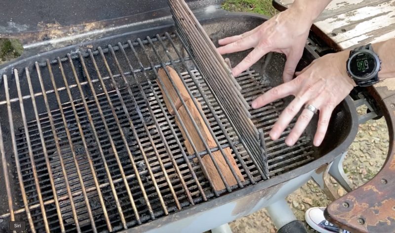 Hands arranging grill grates inside a PK charcoal grill to create a two-zone setup with a chunk of hardwood placed beside the coal bed for indirect heat.